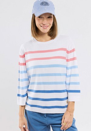 Femme portant un pull blanc avec des rayures horizontales roses et bleues, un pantalon bleu et une casquette rayée bleu et blanc, souriant légèrement.