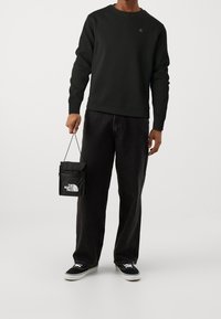 Sweat-shirt noir à col rond et poignets côtelés, associé à un jean noir droit. Tenant un mini-sac noir avec logo. Baskets blanches.