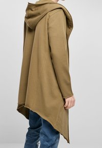 Sudadera sobredimensionada en color caqui con una textura suave, que presenta un dobladillo largo y asimétrico y una capucha profunda, llevada sobre unos jeans azules.