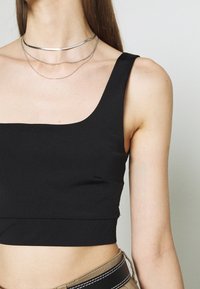 Svart kort topp med fyrkantig halsringning och tjocka axelband, tillverkad av ett mjukt material. Bärs med lager av silverhalsband.