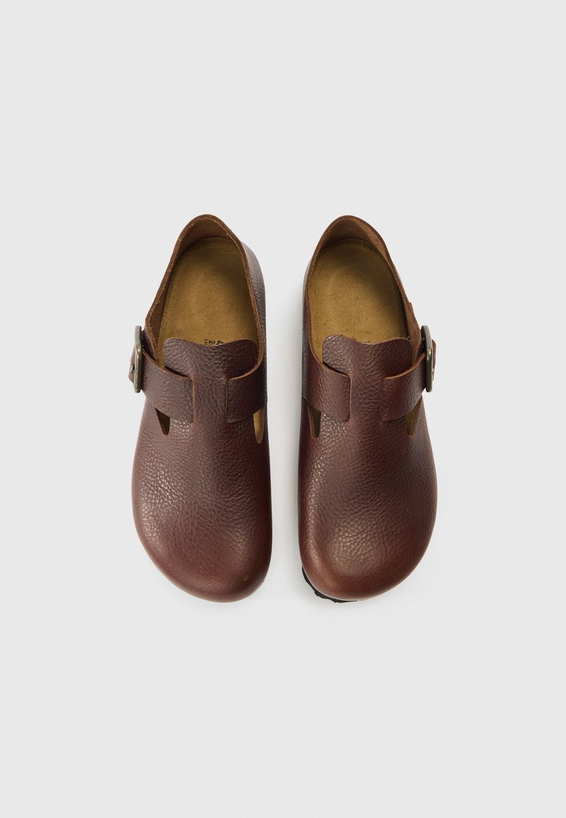 Zoccoli in pelle marrone con finitura testurizzata, forma arrotondata, dettagli traforati e chiusura laterale con fibbia. Plantari morbidi e suole piatte.