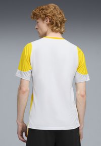 Camisa deportiva para hombre con un cuerpo blanco, mangas amarillas y detalles en gris con patrón en las mangas. Diseño de manga corta y ajustado.