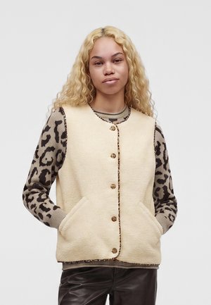 Vrouw met lang krullend blond haar draagt een crèmekleurig vest met knopen over een luipaardprint shirt met lange mouwen en een donkere broek, handen in de vestzakken.