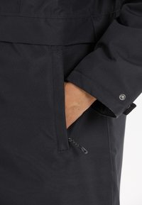 Giacca nera da esterno con una superficie liscia, tasca con zip e dettaglio a pressione sulle maniche. Il materiale è resistente all'acqua e durevole.