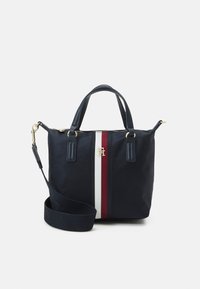 Sac à main bleu marine avec une bande verticale blanche et rouge, logo doré, deux poignées et une bandoulière ajustable sur fond blanc.