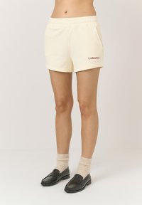 Kremowe szorty z elastycznym pasem i bocznymi kieszeniami. Na dolnej lewej stronie znajduje się czerwony napis "LAMANIA". Gładka faktura.