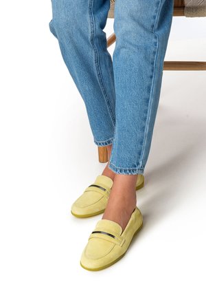 Gele suède loafers met een gladde textuur, voorzien van een zwarte accentstreep, gedragen met lichtblauwe spijkerbroek.