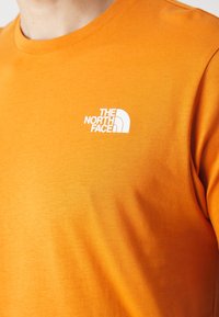 Oranžové bavlněné tričko s bílým logem na levé horní straně. Látka má hladký povrch a klasický kulatý výstřih.