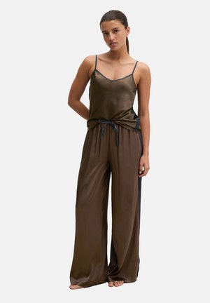 Donna con capelli castani raccolti in una coda di cavallo, indossa pantaloni marroni in raso a gamba larga e un top coordinato con spalline sottili, in piedi a piedi nudi su uno sfondo semplice.