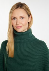Pulover turtleneck rib uitat în verde închis, cu un guler înalt. Prezintă o textură moale și o croială lejeră, purtat cu cercei aurii rotunzi.