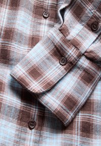 Tissu de chemise en flanelle à carreaux marron et bleu clair avec boutons marron, montrant un poignet de manche et une partie de l'avant avec détails des boutons.