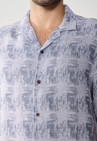 Camisa de manga corta en azul claro y blanco con un patrón texturizado, un bolsillo en el pecho y botones marrones en un cuello relajado.