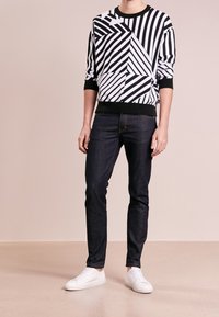 Schwarz-weißer gestreifter Pullover mit geometrischen Mustern, kombiniert mit dunklen Jeans und weißen Sneakers; Modell steht vor einem neutralen Hintergrund.