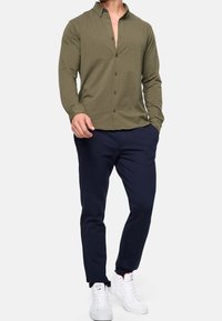 Olive green long-sleeve shirt with a buttoned collar and front buttons, paired with navy trousers and white sneakers.