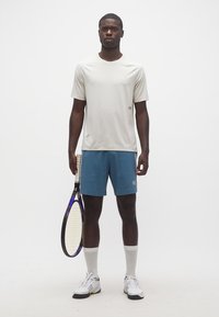 Άνδρας όρθιος που κρατά ρακέτα τένις, φοράει ανοιχτό μπεζ μπλουζάκι, μπλε σορτς, λευκές αθλητικές κάλτσες και λευκά αθλητικά παπούτσια.