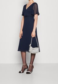 Mörkblå klänning med korta, fladdriga ärmar och v-ringning. Genomskinligt tyg vid ärmarna, kombinerat med en silverfärgad handväska och svarta stilettklackar.