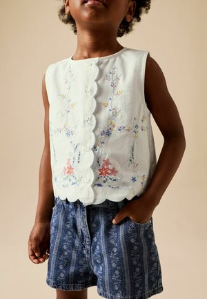 Gyermek fehér ujjatlan blúzban, virágos hímzéssel és hullámos szegéllyel, kék mintás rövidnadrággal, az egyik kezét a zsebében tartva áll.