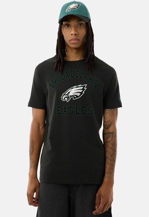 Uomo che indossa una maglietta nera dei Philadelphia Eagles e un cappellino verde degli Eagles, in piedi con le braccia rilassate, espressione neutra.