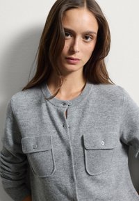 Cardigan en laine gris avec un col à côtes, deux poches avant et des fermetures à boutons. Texture douce avec une finition lisse et des détails subtils.