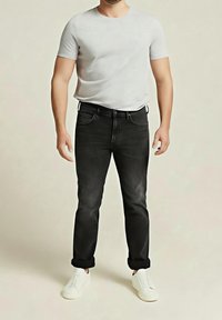Homme portant un t-shirt blanc uni, un jean noir avec ourlets retroussés et des baskets blanches, debout devant un fond neutre.