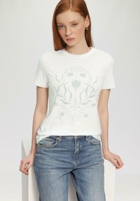 Witte katoenen t-shirt met groene botanische afbeelding en de tekst "DANKBAAR VOOR DE SCHONE DINGEN." Gecombineerd met blauwe denim jeans, relaxte pasvorm.