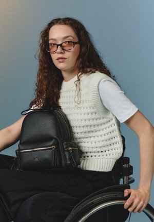 Must must nahkbackpack topelt tõmblukkude, ümmarguse kuju ja kuldsete aksessuaaridega. Mudel valges kampsunis istub ratastoolis.