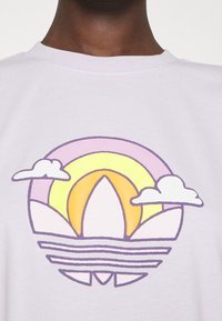 Ljuslila bomullströja med en grafisk design som har en regnbåge och moln i gult, rosa och vitt, placerad över lager av former.