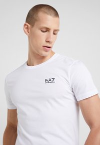 Balts īsām piedurknēm T-krekls no kokvilnas, ar mazu melnu logotipu kreisajā krūtīs. Klasisks apaļas kakla dizains ar gludu tekstūru.