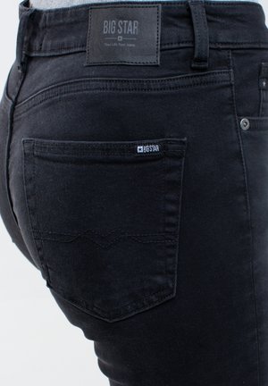 Melnas džinsu bikses ar tuvplānu aizmugurējai kabatai, kurā ir smalks diegu šūjums un neliels logotipa uzlīme tuvu josta. 