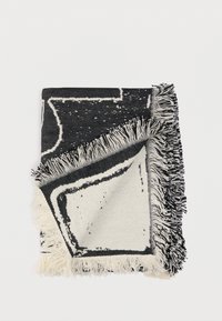 Μαύρη και λευκή υφαντή κουβέρτα με κρόσσια στις άκρες, που παρουσιάζει γεωμετρικό μοτίβο και απαλή υφή. Σερβιρισμένη διπλωμένη και μερικώς ορατή.