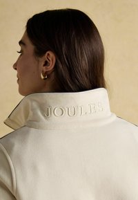 Kremowa kurtka z tkaniny z wysokim kołnierzem z wytłoczonym logo "JOULES", wykończona ciemnym obszyciem na brzegach.