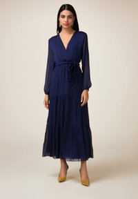Robe maxi bleu marine avec un décolleté en V, des manches longues et une ceinture à la taille. Présente des volants superposés à l'ourlet et une texture de tissu transparent.