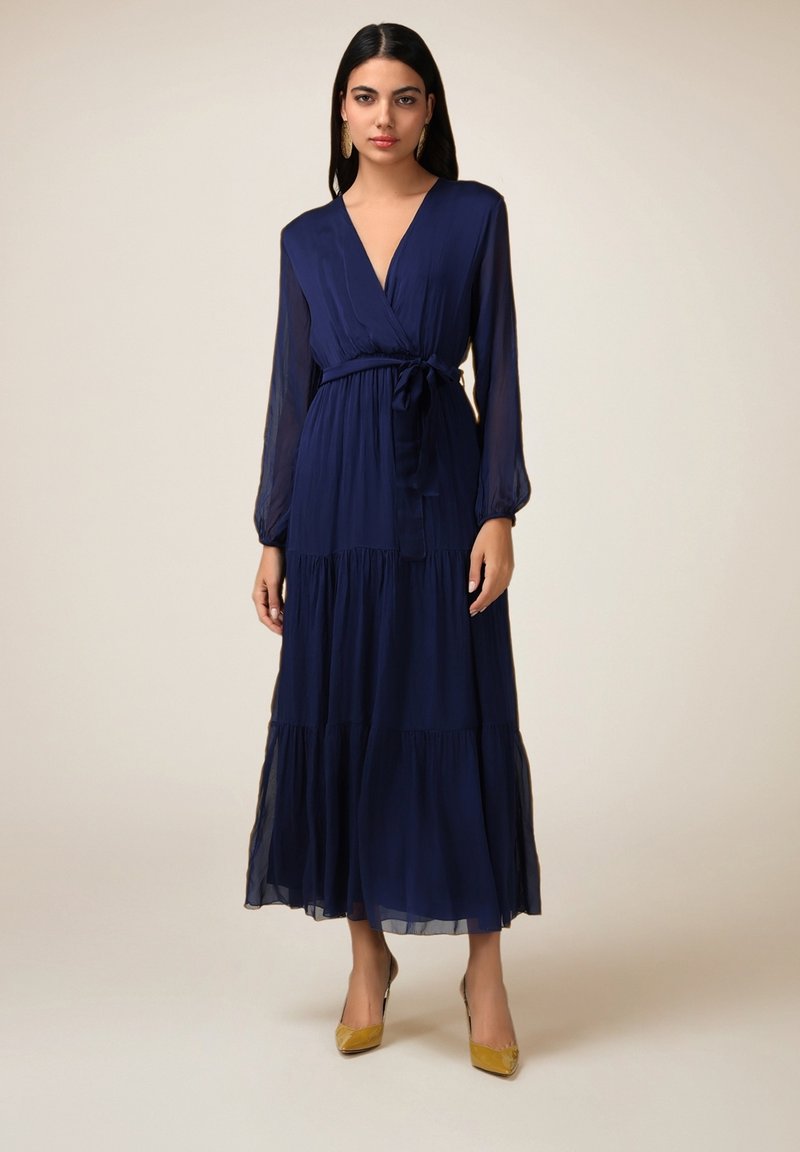 Robe maxi bleu marine avec un décolleté en V, des manches longues et une ceinture à la taille. Présente des volants superposés à l'ourlet et une texture de tissu transparent.
