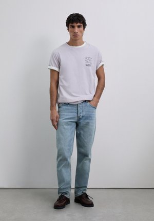 Tânăr stând cu o mână în buzunar, îmbrăcat cu un tricou lavandă cu mânecile rulotate, blugi ușor albaștri și pantofi din piele maro, în fața unui zid gri.