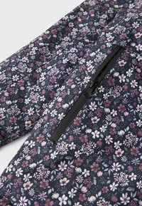 Tumesinise taustaga lillemustriga kangas, millel on väikesed roosad, valged ja rohelised lilled; sisaldab musta, tõmblukuga taskut, millel on sile viimistlus.