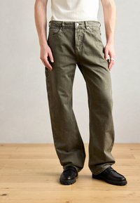 Olive green, loose-fitting pants with a five-pocket design, featuring a button fly and visible stitching details, worn with black shoes.