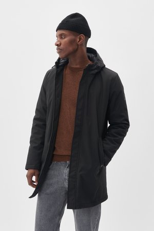 Parka negra impermeable con cierre de cremallera, capucha y dos bolsillos laterales. Se lleva sobre un suéter marrón y jeans grises. Textura suave.