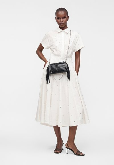 Femme portant une robe midi blanche à manches courtes avec de petits pois, des sandales à talons noires et un sac en cuir noir porté en bandoulière avec des franges.