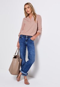 Chemise henley rose clair, jeans bleus, sac à main en cuir beige et sandales à brides argentées. Tenue décontractée avec une coupe ample et des textures douces.