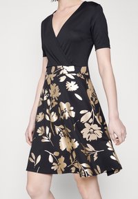 Robe noire portefeuille à manches courtes ; corsage en tissu lisse, jupe à imprimé floral doré ; silhouette en A et longueur au genou.