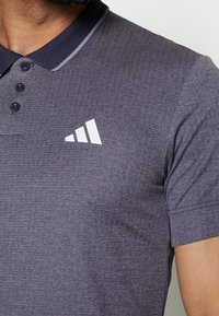 Polo en tissu texturé gris foncé, avec une patte de boutonnage à deux boutons et un logo blanc sur la poitrine. Le col est bordé d'une finition contrastante.