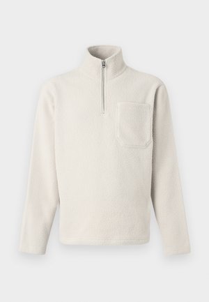 Jasno beżowy polarowy sweter z wysokim kołnierzem, zapinany na pół-zamek, z kieszenią na piersi. Tekstura jest miękka i puszysta.