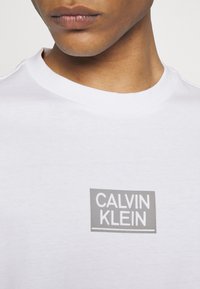 Camiseta de algodón blanca con un logo rectangular gris con el texto "CALVIN KLEIN" en el centro. Cuello redondo y textura suave.