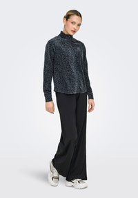 Γυναίκα που φοράει ένα σκουρόχρωμο μπουφάν με φερμουάρ και σχέδια, φαρδιά μαύρη παντελόνα και λευκά χοντρά αθλητικά παπούτσια, στεκόμενη με ελαφρύ σκύψιμο σε απλό φόντο.