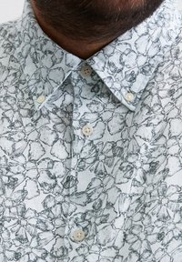 Hellblaues Leinenhemd mit einem detaillierten schwarzen Blumenmuster, einem Button-Down-Kragen und zwei cremefarbenen Knöpfen in der Nähe des Kragens.