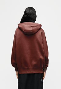 Άτομο με μακριά σκούρα μαλλιά, ντυμένο με φαρδιά, σκουρόκόκκινη φούτερ μπλούζα με κουκούλα και μαύρο παντελόνι, στέκεται γυρισμένο με την πλάτη προς ένα απλό φωτεινό φόντο.