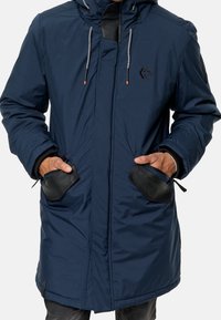 Parka bleu marine en tissu imperméable, dotée d'une capuche, de poches zippées avec bordure noire et de cordons ajustables.