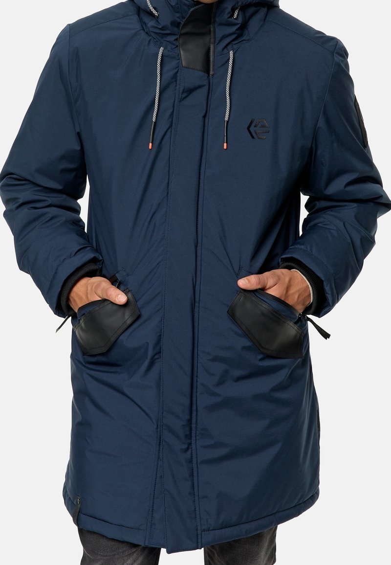 Parka bleu marine en tissu imperméable, dotée d'une capuche, de poches zippées avec bordure noire et de cordons ajustables.