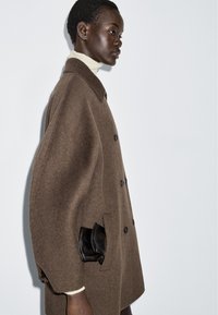 Brun ullrock med knappfram, överdimensionerad design och en unik detalj med svart läderficka på sidan.