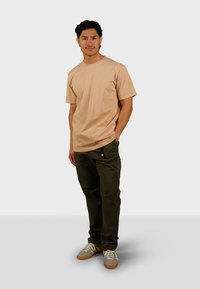 Beige T-shirt met korte mouwen, ronde halslijn en een klein logo. Olijfgroene broek met zakken en een taps toelopende pasvorm. Grijs en tan sneakers.